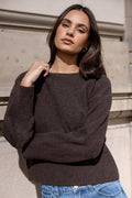 Woman wearing a dark brown sweater and blue jeans standing against a stone wall.