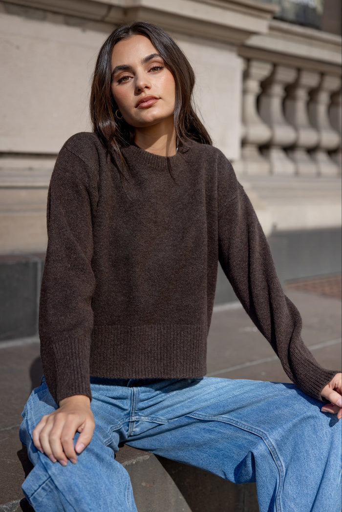 Woman wearing a brown sweater and blue jeans sitting on steps.