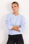 A woman wearing a light periwinkle blue ribbed knit sweater with black pants, standing against a plain white studio background.