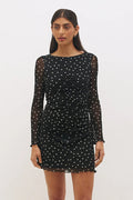 A woman modeling a black long-sleeve mini dress with white polka dots and sheer mesh sleeves, standing against a plain light gray studio background.