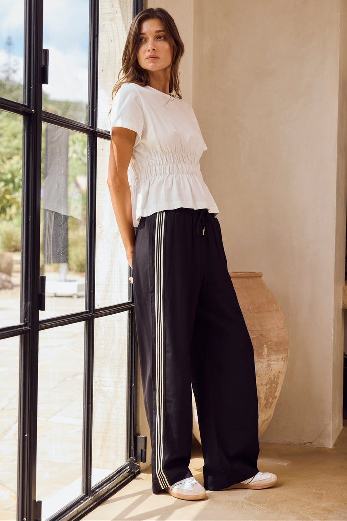 Woman wearing a white top and black pants with white stripes standing near a glass door.