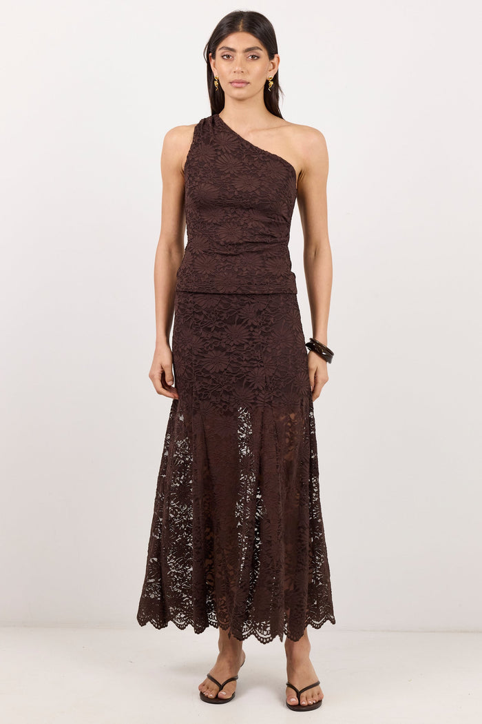 A woman modeling a dark brown one-shoulder lace maxi dress with a fitted bodice and flared skirt, paired with black strappy sandals, against a plain white studio background.