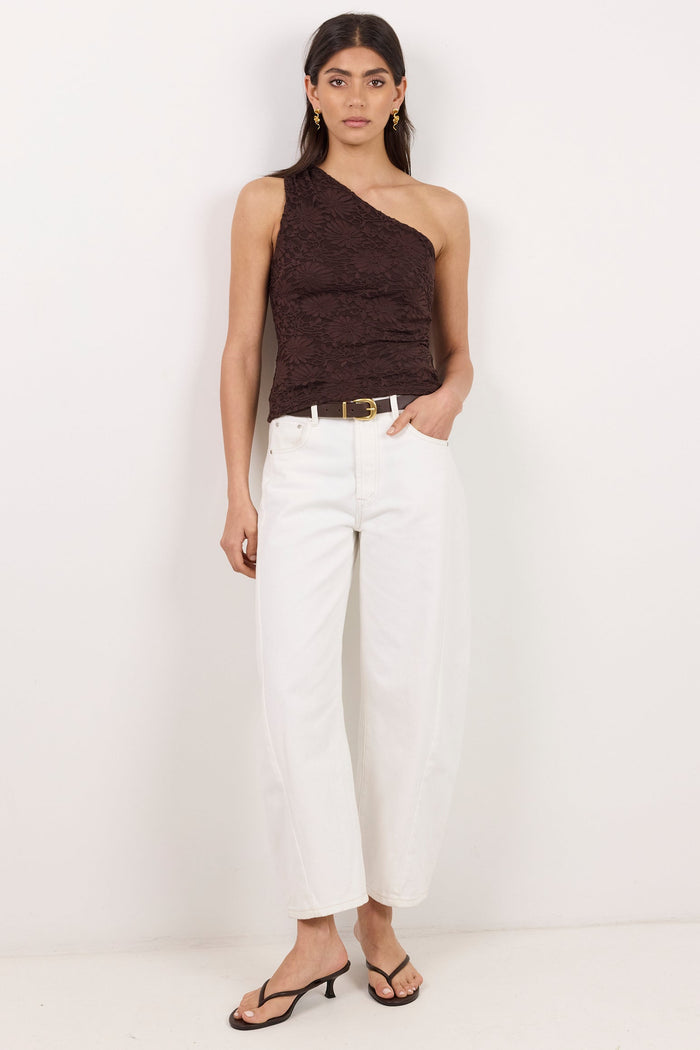 A woman stands against a plain white studio background wearing a dark brown one-shoulder textured top, white cropped pants, a thin gold belt, and black heeled sandals.
