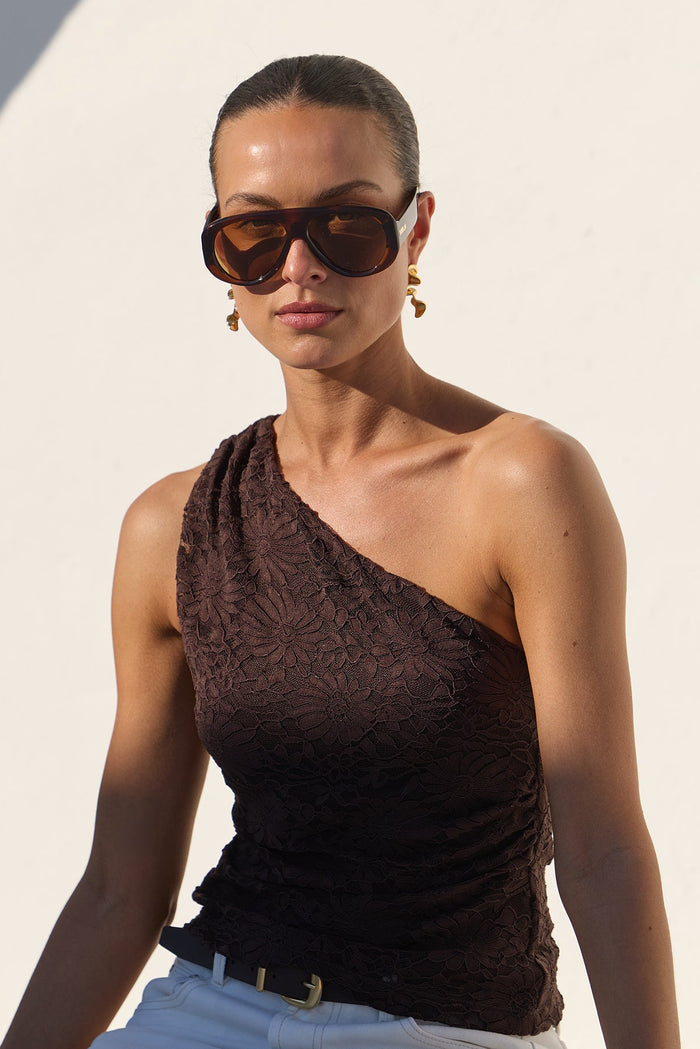 A woman wearing a dark brown one-shoulder textured top, white trousers, and sunglasses sits on a white architectural ledge in bright sunlight.