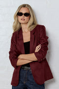 Person wearing a burgundy blazer and sunglasses against a white brick wall