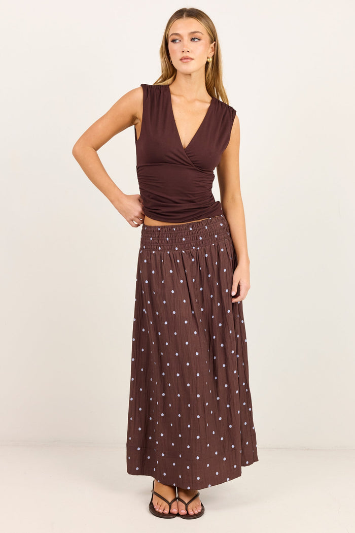 A woman modeling a sleeveless dark brown wrap-style top and a matching dark brown maxi skirt with small white polka dots, standing against a plain off-white studio background.