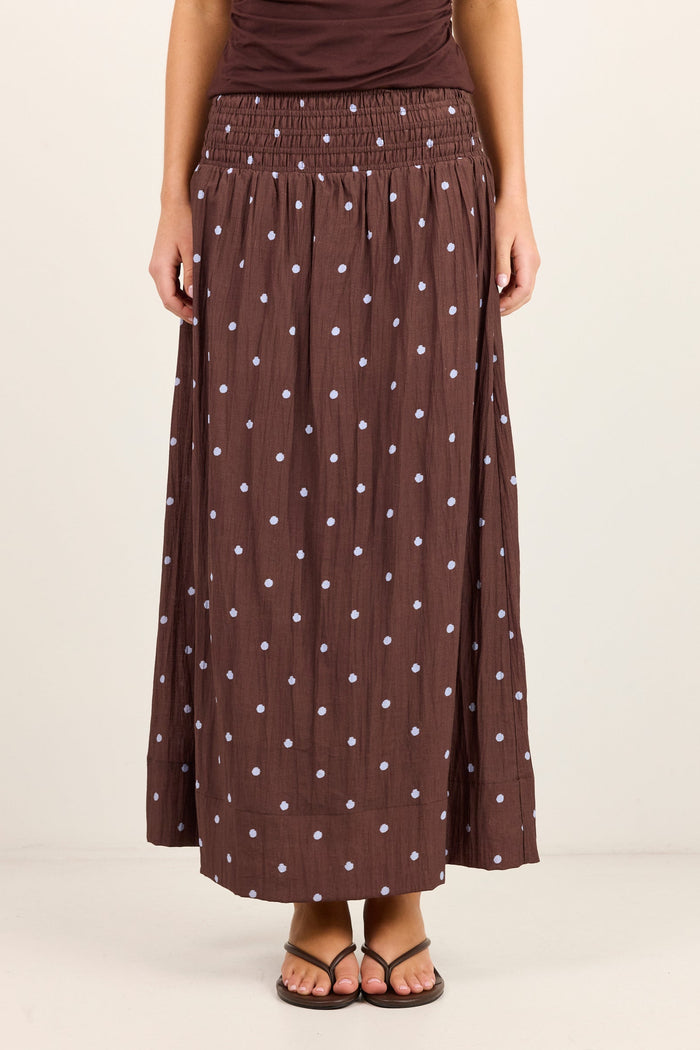 A model wearing a brown pleated maxi skirt with a white polka dot pattern, paired with a matching brown top and brown sandals, against a plain off-white studio background.