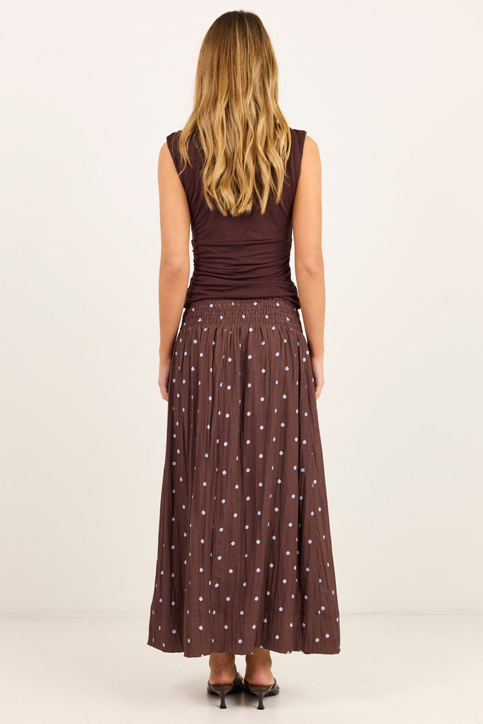 A woman viewed from the back wearing a sleeveless dark brown top and a matching long pleated skirt with small white polka dots, standing against a plain light studio background.