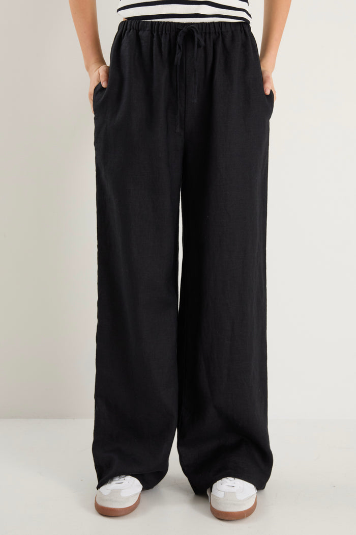 Woman wearing black wide-leg linen pants with an elasticated waistband, styled with a black and white striped tank and white sneakers.