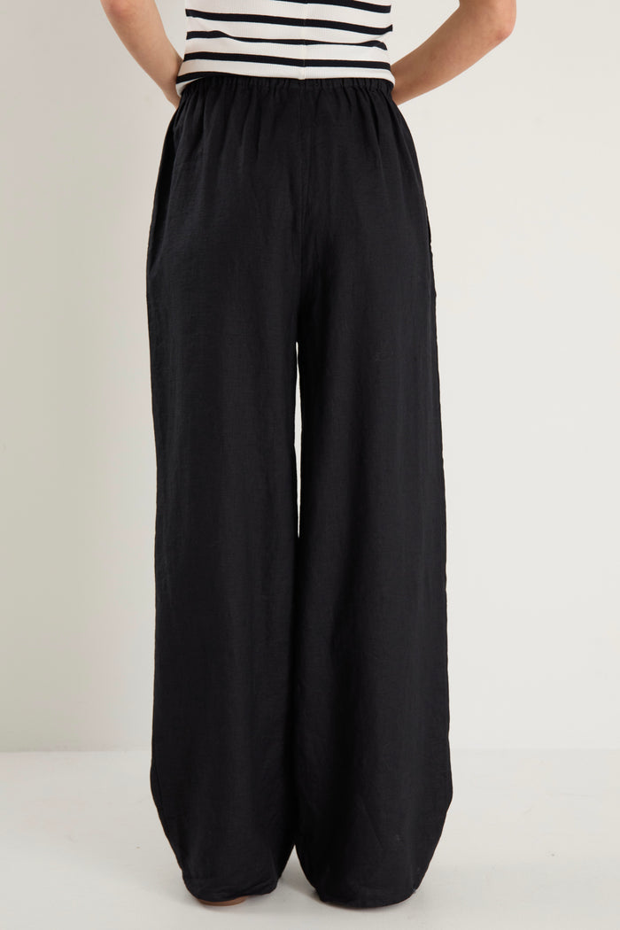 Woman wearing black wide-leg linen pants with an elasticated waistband, styled with a black and white striped tank and white sneakers.