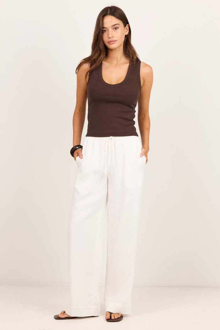 A woman modeling a dark brown ribbed tank top and white wide-leg linen pants against a clean, off-white studio background.