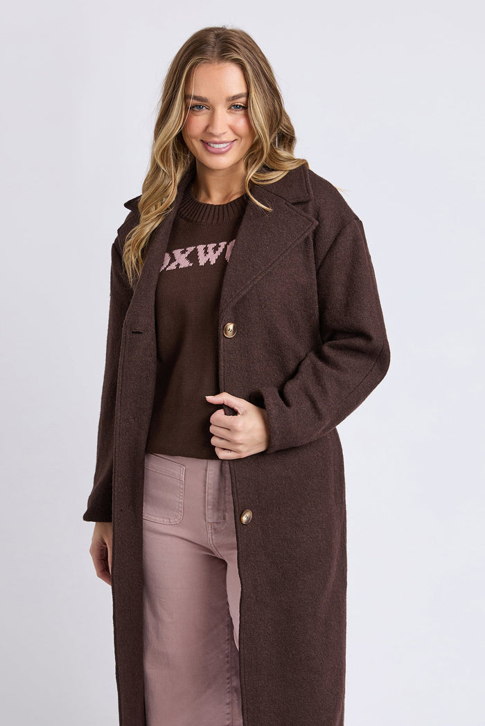 Woman wearing a brown coat over a brown sweater with visible text on a light gray background