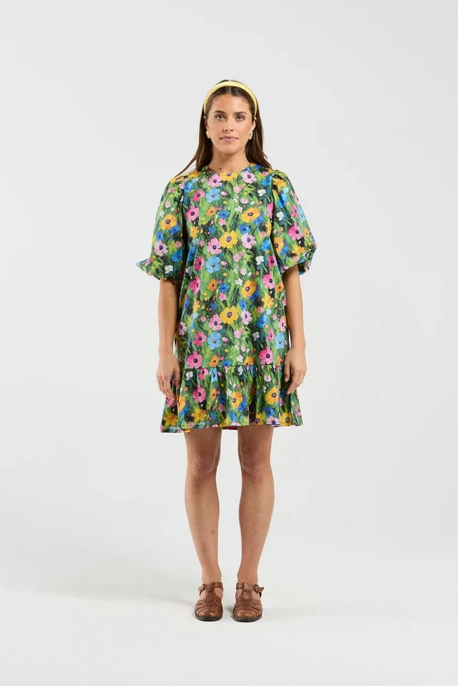 A woman stands against a light gray studio background wearing a short-sleeve tiered dress with a colorful floral print, brown flat shoes, and a yellow headband.