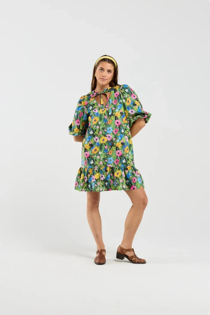 A woman modeling a short green floral dress with puff sleeves, a tie neckline, and a ruffled hem, paired with a yellow headband and brown sandals, against a plain light gray studio background.