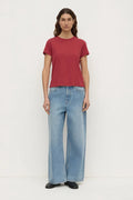 A woman stands against a plain white studio background wearing a short-sleeve crew-neck t-shirt in a muted brick red color, paired with light-wash wide-leg blue jeans and black shoes.