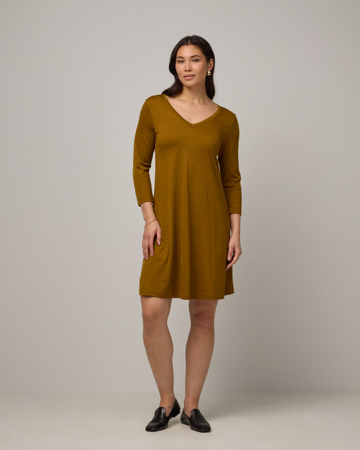 Model wearing Willow Swing Dress in Spiced Pear, a dark mustard color. Willow Swing Dress is a merino wool long sleeve, v-neck, knee length swing dress with pockets available in sizes XS-3X Regular, XS-3X Long.