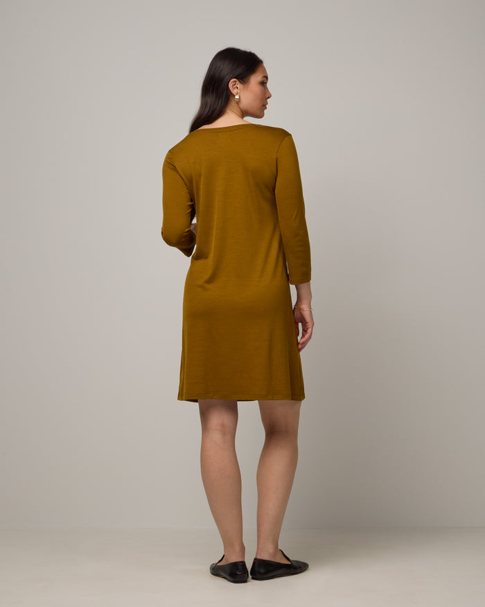 Model wearing Willow Swing Dress in Spiced Pear, a dark mustard color. Willow Swing Dress is a merino wool long sleeve, v-neck, knee length swing dress with pockets available in sizes XS-3X Regular, XS-3X Long.