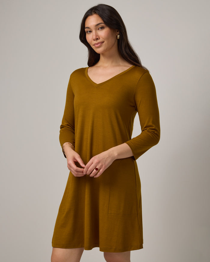 Model wearing Willow Swing Dress in Spiced Pear, a dark mustard color. Willow Swing Dress is a merino wool long sleeve, v-neck, knee length swing dress with pockets available in sizes XS-3X Regular, XS-3X Long.