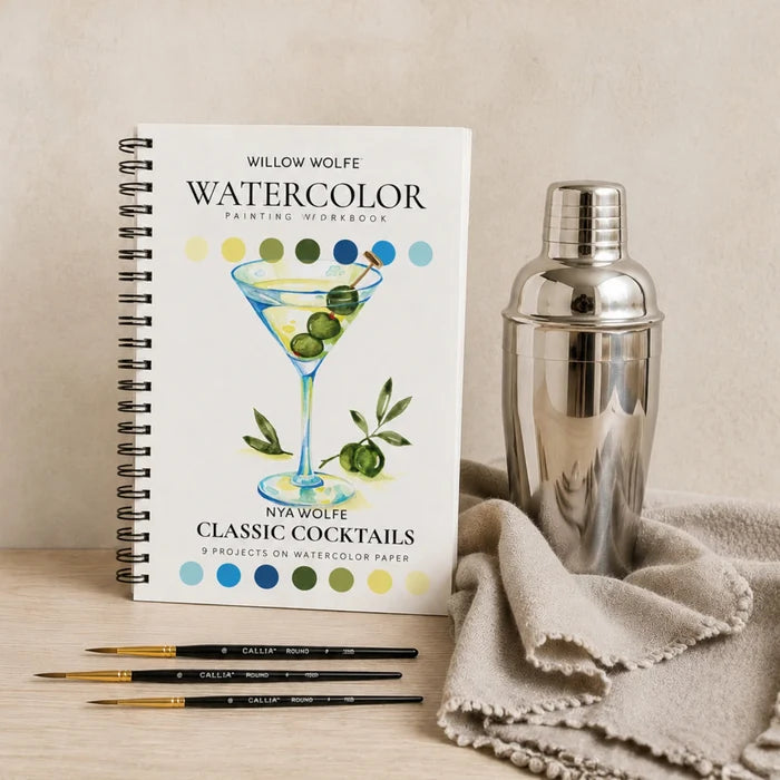 A spiral-bound watercolor painting workbook titled 'Willow Wolfe Watercolor Painting Workbook' featuring a martini glass illustration, styled on a tabletop next to a silver cocktail shaker, a beige cloth, and three paintbrushes.