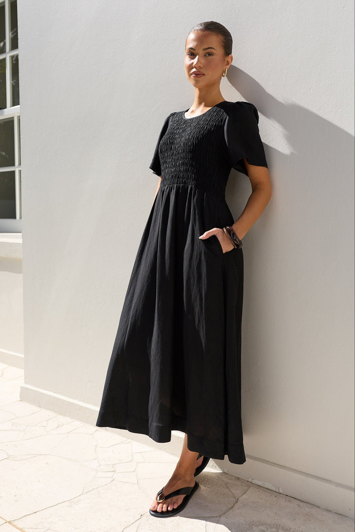 A woman wearing a black midi dress with a smocked bodice and short puff sleeves, leaning against a white stucco wall outdoors.