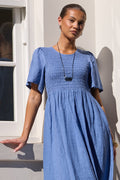 A woman wearing a smocked blue linen maxi dress with short puff sleeves, standing against a white stucco wall with a window on the left.