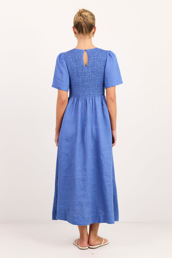 A woman viewed from the back wearing a blue midi dress with short puff sleeves, a smocked bodice, and a keyhole back detail, standing against a plain white studio background.