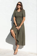 A woman wearing an olive green midi dress and sunglasses stands next to a swimming pool in front of a white wall.
