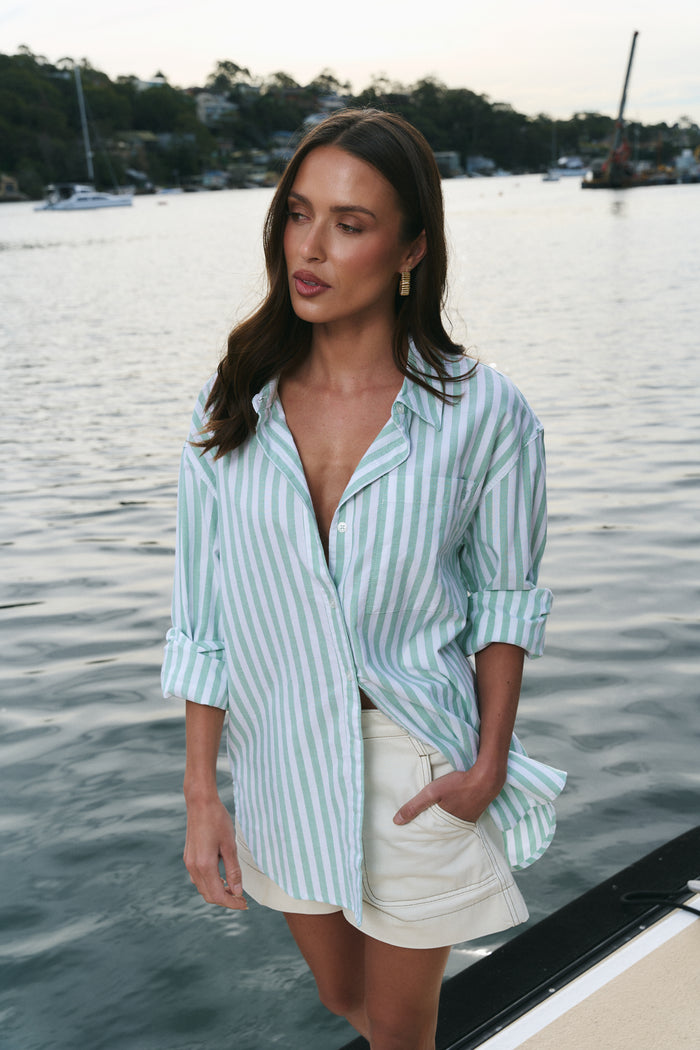 Woman wearing a light blue striped shirt and beige shorts by a waterfront.