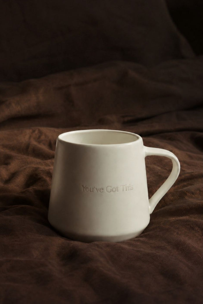 A matte white ceramic mug with the phrase 'You've Got This' printed in a subtle, tonal font on the front, resting on a dark brown textured fabric background.