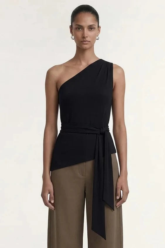 A model stands against a plain light background wearing a one-shoulder black wrap top and wide-leg taupe-brown pants with black sandals.