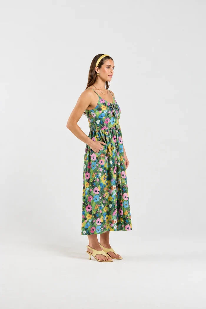 A woman modeling a sleeveless midi dress with a colorful floral print on a green base, paired with a yellow headband and light yellow sandals, against a plain white studio background.