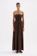A woman modeling a strapless, smocked-bust maxi dress in a solid dark brown color, standing against a plain white studio background.