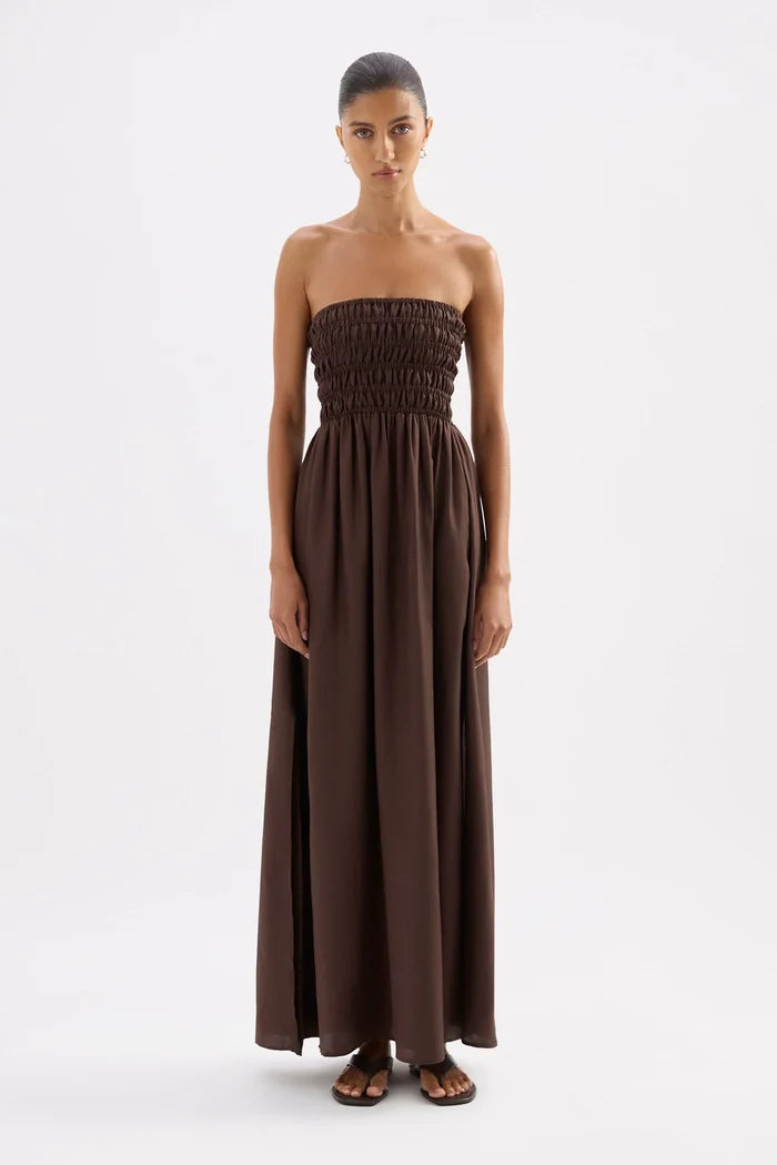 A woman modeling a strapless, smocked-bust maxi dress in a solid dark brown color, standing against a plain white studio background.
