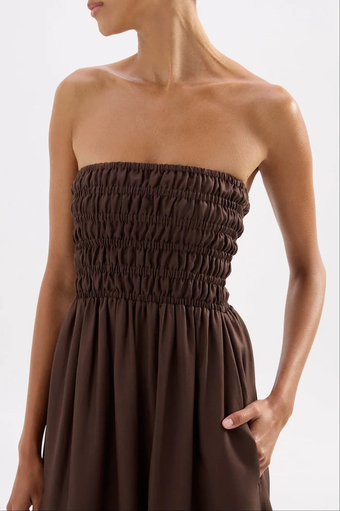A close-up of a model wearing a dark brown strapless smocked dress against a light gray studio background.