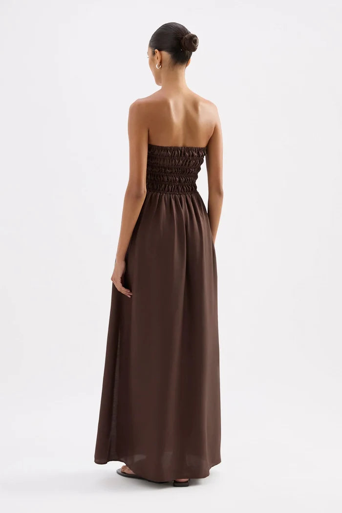 A woman viewed from the back wearing a strapless dark brown maxi dress with a smocked bodice and flowing skirt, standing against a plain light gray studio background.